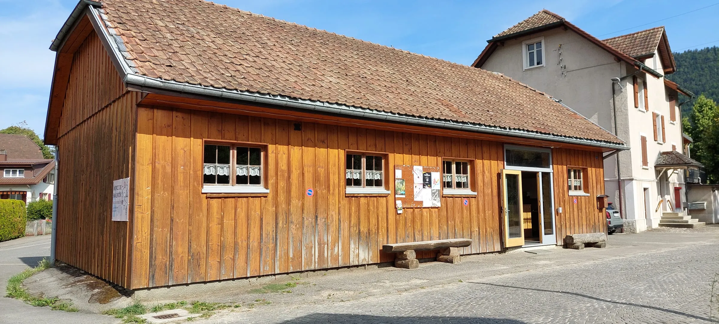 La façade en bois et sa toiture en tuiles, vue de biais depuis la place